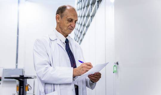 A man in a lab coat writes notes on a piece of paper standing in a pharmaceutical storage depot.