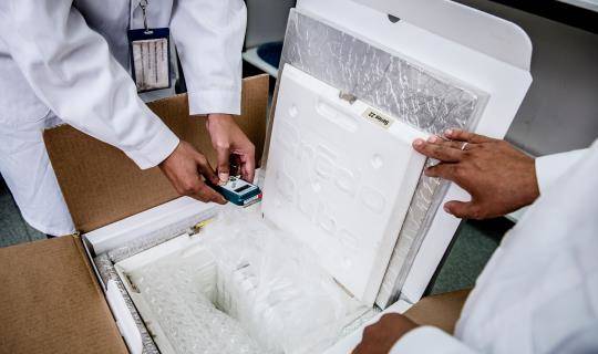 Two people in lab coats take a reading from a digital monitor inside an insulated package.