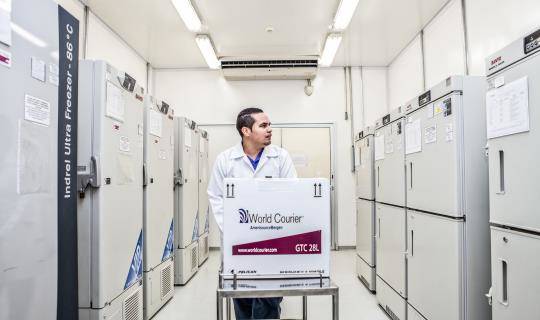 A man pushes a box on a cart down a wide aisle of storage lockers in a pharmaceutical storage depot.