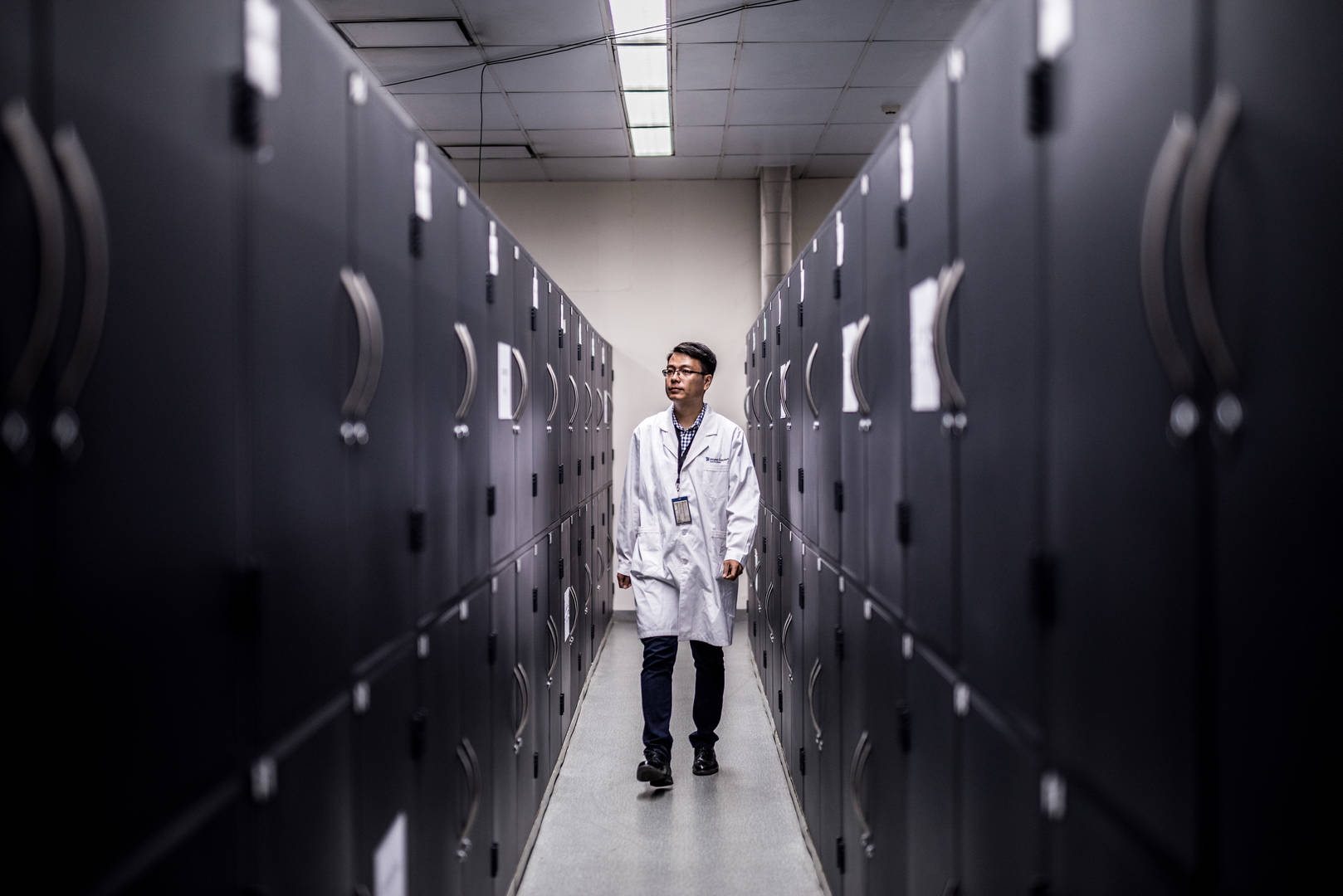 A man in a lab coat walks down a narrow aisle of lockers in a storage depot.