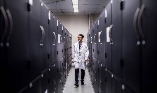 A man in a lab coat walks down a narrow aisle of lockers in a storage depot.