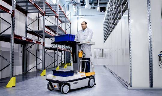 A man drives a forklift holding a small bucket down a wide aisle of shelves in a storage depot.