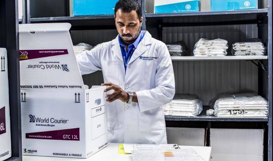 A World Courier associate in a lab coat packs a box for shipment surrounded by shelves.