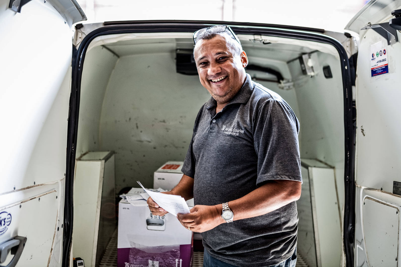A smiling World Courier driver stands in front of his van, which is open and shows packages inside.