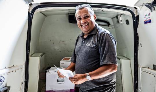 A smiling World Courier driver stands in front of his van, which is open and shows packages inside.
